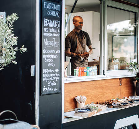 Kock som serverar mat från Egastronomis matvagn, med meny skriven på griffeltavla och upplagda rätter i luckan.